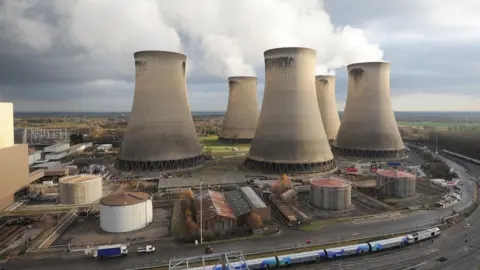 Five large power station chimneys with white smoke coming out of them stand surrounded by much smaller industrial buildings. A road circles around the site. Behind the site there are fields and trees.