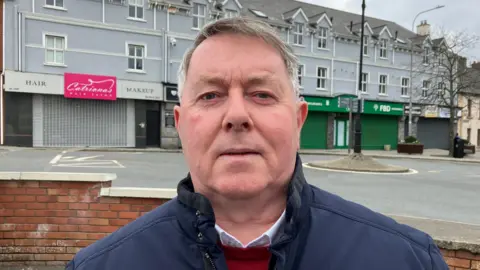 A man with short gray hair looks directly into the camera. He is wearing a navy coat, a white shirt and a red jumper. Behind him is a terraced row of shops and businesses. 