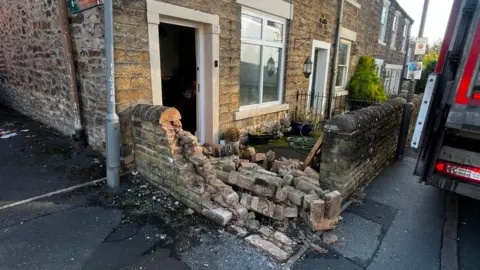 A front garden wall which has been knocked down - there are bricks all over the front yard and to the right there is a lorry parked on the pavement. 