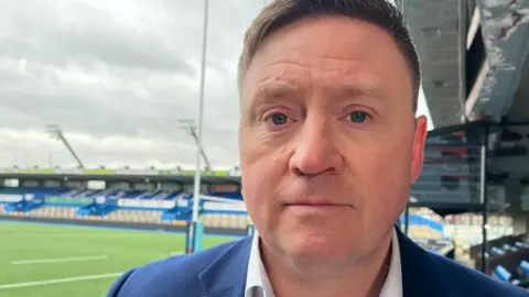 Rhodri, yang memiliki rambut hitam pendek, mengenakan blazer biru tua dan kemeja putih. Di latar belakang adalah stadion kosong dan nada Cardiff Arms Park. 