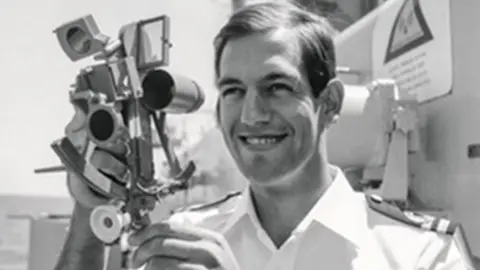 A black and white photo shows a man in white naval uniform, holding an old-fashioned camera. He is smiling as he looks just to the side of the camera taking his picture. He appears to be standing on board a ship and a small section of sea can be seen behind him.