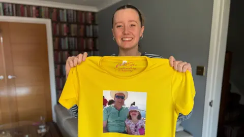 Carys Walsh holds up her yellow race t-shirt, which has a photo of her with her dad Rod on the front. She is standing in a living room. There are brown double doors in the background. There is also a shelf with books.
