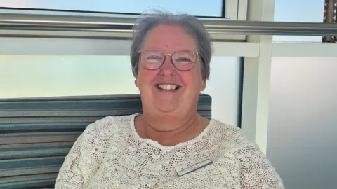 A woman in a white top is smiling at the camera. She is wearing glasses and a window is directly behind her.