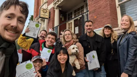 FoBGWMC A group of men and women smiling at the camera holding up A4 pieces of paper that say 'Save BGWMC'. One person is holding a fluffy white dog in the middle of the group. 