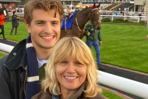 Scott Family A woman with shoulder-length blonde hair and a man with short brown hair stand by a white rail at a racecourse with a brown horse being led by the reins in the ring behind them. Both people are smiling at the camera. 