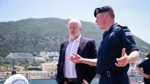PA Media Two men, on the left is Al Carns wearing a dark suit jacket, trousers and brown belt, with a white shirt underneath. To his right is a man wearing a military uniform. They are pictured talking on an outside deck of a boat. In the distance, behind them there is a built up residential along the water and a mountain. 