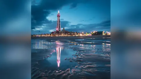 Quintin Lake A view of Blackpool Tower and the seafront at night, with reflections in the mud at the foreground of the photo.