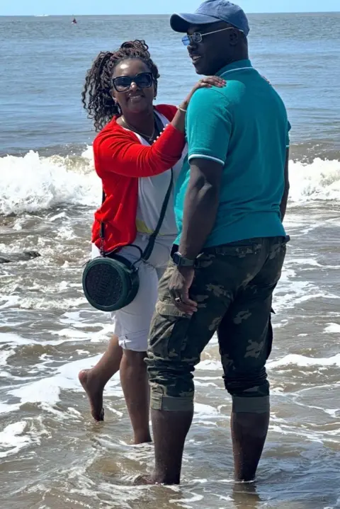 Emertha Uwanyirigira Emertha Uwanyirigira and her husband George Libon standing ankle deep in the sea. Emertha is wearing white shorts and a white T-shirt with a red cardigan and a green circular hand bag sitting on her hip. She has dark sunglasses on and is looking at the camera with her hands on her husband's shoulders. George has his back to the camera but is turning his face to the left. He is wearing camouflage trousers that are rolled up the knee and a turquoise polo T-shirt with blue-lensed sunglasses and a blue baseball cap.
