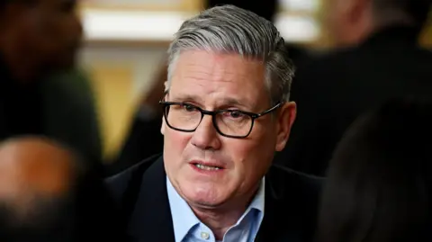 Keir Starmer, in an open neck shirt and suit jacket, looking pensive