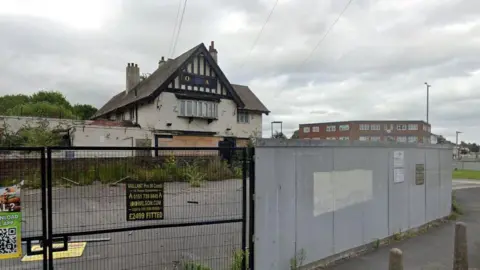 A deteriorated pub sits behind a black fenced gate and grey painted wall