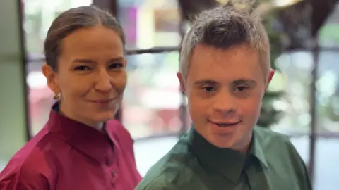 TEAM DOMENICA The image shows two people smiling, a woman on the left and a man on the right. The man is wearing a green shirt and the woman, a red one. They are both smiling and behind them are blurred windows.