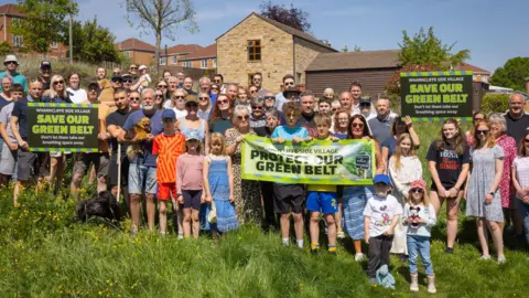 Dozens of people are stood in a green field, with houses in the background. They are holding placards and banners saying "protect our green belt"