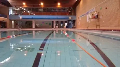 A swimming pool with blue tiles, black painted lane markers and orange rope lane dividers. It is housed inside yellow-brick walls with a viewing area at the far end.