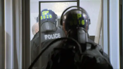 Two police officers wearing black tactical clothing and helmets with yellow writing, carrying riot shields.