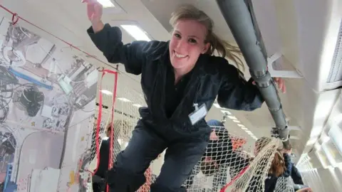 Susan Charlesworth has long blonde hair and is wearing a green jumpsuit. She is in an anti-gravity plane and floating near the ceiling.