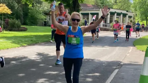 Sue Harley Ms Harley is raising both arms as she takes part in a race. She is wearing a blue vest top and dark leggings with her name and race number on a green tab on her chest. Her blonde hair is tied back and she has black shades on.