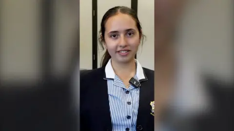 A school girl with hair tied back and a blue and white striped shirt under a uniform blazer.