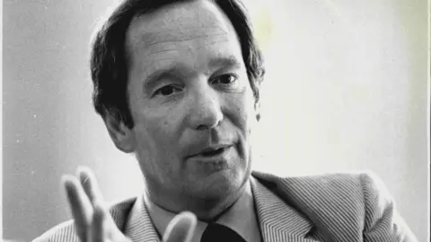 Getty Images Close up black and white image of broadcaster Michael Buerk. He looks like he is speaking and is gesturing with his right hand. He is wearing a striped suit jacket with a shirt and tie.