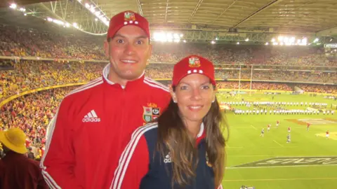 Matthew Miles An image of a man and a woman in the stands of a sports stadium. The man is wearing a red track suit and red baseball cap and the woman is wearing red and blue tracksuit and a red baseball cap. In the background can be seen a stadium with a capacity crowd.
