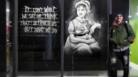 Ryan Stanikk Man with green jacket with his hands in his pockets standing outside by a window decorated with an image of Jane Austen sprayed with fake snow.