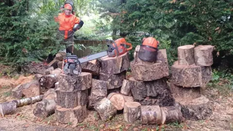 Fiona Read A man with a high vis jacket and safety helmet stands behind a big pile of logs on which two chainsaws sit.