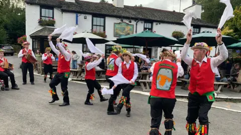 Supplied Furness Morris Dancers performing in Elterwater in July 2023. They have formed a line and a are waving white cloths in the air as three members at the back play some music. They are dressed in black trousers, white shirts and red waistcoats.