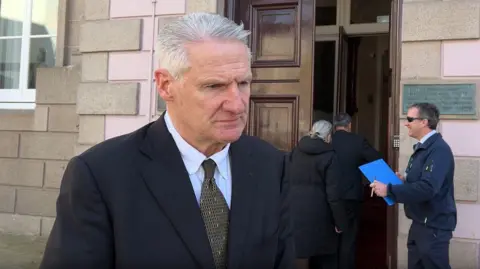 BBC Health Minister Tom Binet looks to the side of the camera as he's being interviewed outside the States Assembly. He has a dark blue jacket on and a light blue shirt with a black and patterned tie. He has short white hair.
