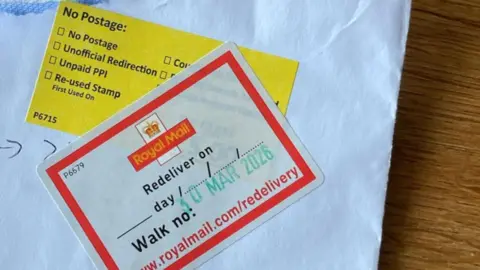 Supplied The corner of an opened white envelope, onto which two stickers have been stuck. A yellow sticker says "no postage", while another - placed over the top of the yellow one - has Royal Mail's logo on it, with "redeliver on 30 Mar 2026" also written.