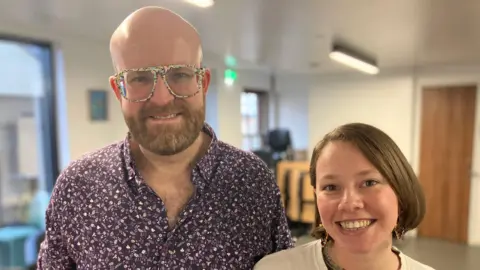A bald man with multicoloured glasses and a purple patterned shirt. Next to him is a woman with a bob haircut in a white t-shirt. Both are looking towards the camera smiling.
