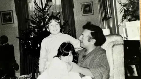 PA Media A black and white photo showing a young boy and girl sitting on their father's knee in an armchair in front of a Christmas tree. The three are smiling.