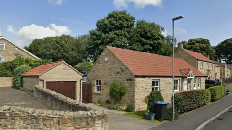 Close-up shot of a small bungalow with a large tree behind it. The bungalow has light-coloured bricks and an orange-brown slate roof.