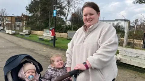 A woman is standing with two children. One is in a buggy and is wearing a light-coloured wooly hat. There are wooden benches in the background.