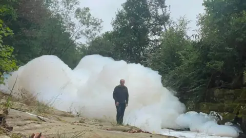 Craig Hannah Large white clumps of foam in a river. A man stands next to the foam and it is taller than him by a significant amount.