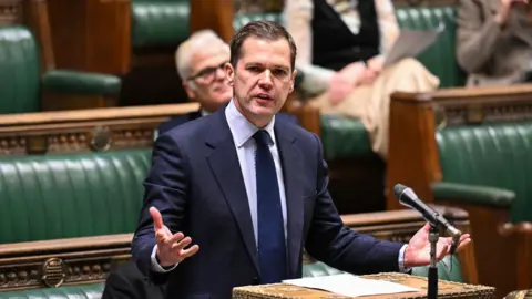 PA Media A man in a navy blue jacket and matching tie stands in front of a microphone in the House of Commons, gesturing with his hands as he addresses other MPs.
