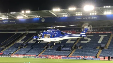 Pete White Helikopter G-VSKP dengan eksterior biru putih melayang di atas lapangan di dalam stadion sepak bola yang kursinya berwarna biru.