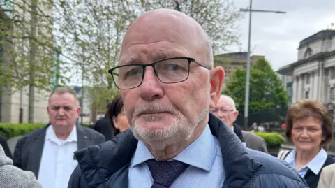 A bald man with grey stubble, looking at the camera. He is wearing a blue shirt, navy tie and navy coat. There is a group of people behind him. 