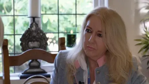 Janis James, who has long blonde hair, is sitting on a wooden chair wearing a blue and pink jacket. She has a window behind her with a candle stick on the window sill. 