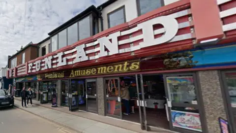 The image shows the exterior of a two-storey seaside amusement arcade with a long, continuous frontage facing the street. The upper floor has light-coloured walls and several rectangular windows, some of which are grouped together in a bay section. The roofline is flat. A large, bold sign stretches across the width of the building. It reads FUNLAND in oversized white block letters mounted on a red background with horizontal striping. Beneath this another sign reads Funland Amusements in gold lettering on a darker red band.
