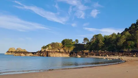 BBC Weather Watchers/Hedi Gorey in Jersey. A sandy beach with blue clear water. The sky is blue with white clouds. In the background are trees. 