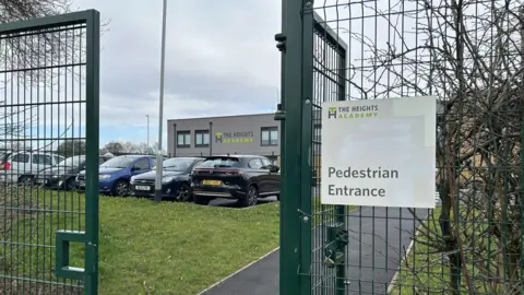 Heights Academy in Smith's wood. A pedestrian entrance hangs on a green metal gate at an entrance of the building. The school building is in the background and a car park can also be seen. 