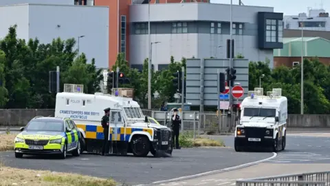 BBC Two armoured police vehicles and a police car parked nearby. Two police officers are standing beside one of the armoured vehicles which has paint splattered on it.
