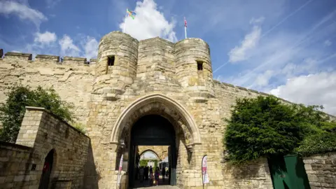 Imposing, high stone castle walls and a large gatehouse with an arched entrance and two circular towers above it topped with flagpoles flying the flags of Lincolnshire and the Union. People can be seen walking under the archway. The sky is blue.