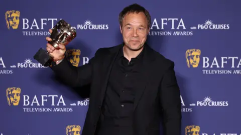 EPA Stephen Graham wears a black suit and shit and is smiling holding a gold BAFTA award. He is stood in front of a purple backdrop which is covered in the BAFTA logo. He has short dark hair