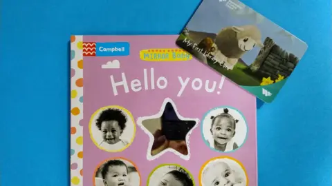 A children's book titled Hello you! It has a pink front cover with five images of babies' faces along with a star. One of the library cards being given out by Westmorland and Furness Council is placed alongside the book. It says "My First Library Card" and has an illustration of a sheep.