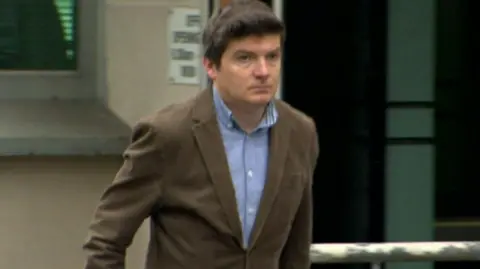 McMonagle walking outside a building. He has short brown hair, and he's wearing a brown suit with a blue shirt.