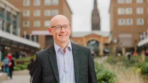 Coventry City Council Martin Reeves, with glasses, smiling with Coventry city centre in the background