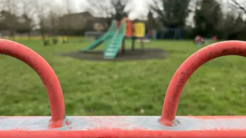 Emma Baugh/BBC A children's play area. Metal railings in the foreground are visible, with swings and a slide in the background