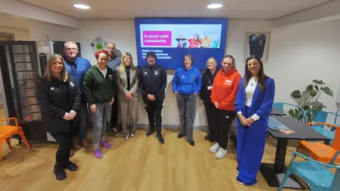 North East Lincolnshire Council A group of ten people stood in a room in front of a screen. The floor is wooden, the walls are white and there are black tables and blue and orange chairs in the background. 