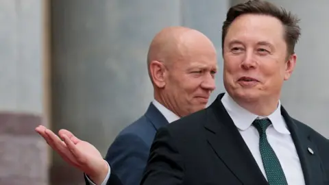 Elon Musk leaving a building in China, smirking and gesturing with his hand while wearing a black suit jacket, white collared shirt and dark green tie.