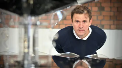 Bob Pitchford A man looks at the camera with a serious expression, through the gap in a metal sculpture. He has short brown hair and is bending forwards. He is wearing a navy thin jumper and a white collared shirt. 